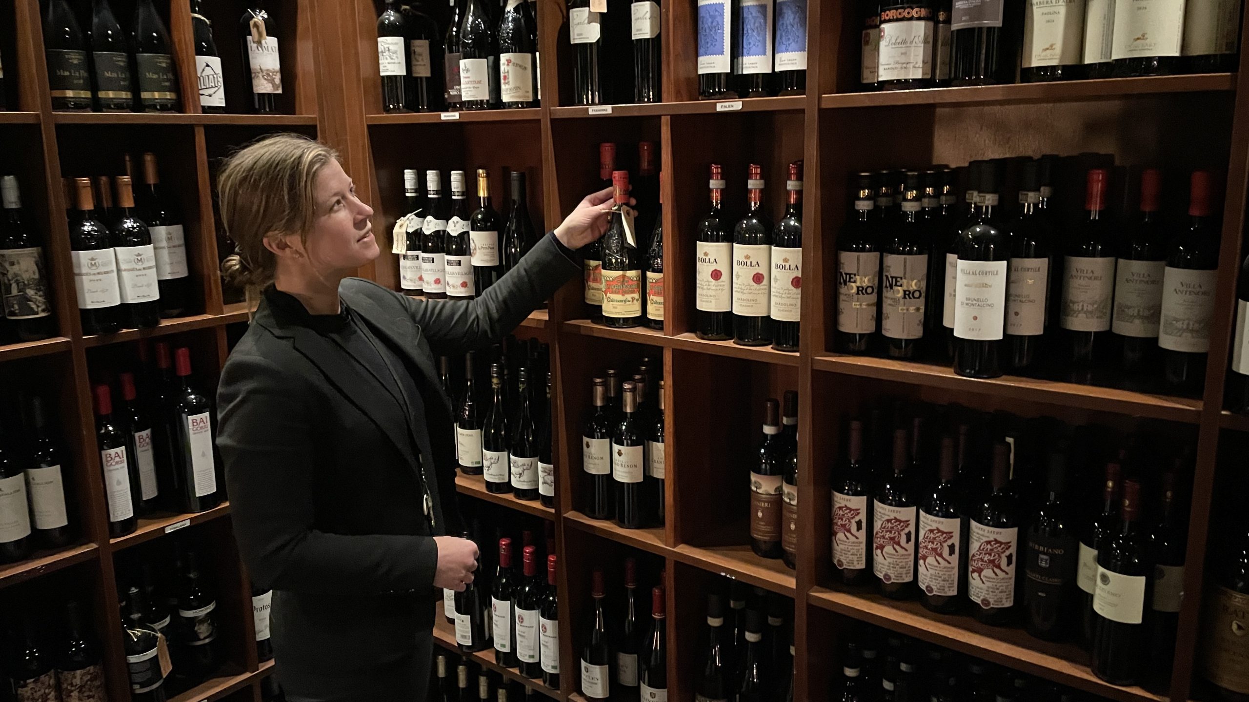 Kvinna i kavaj står i dunkelt upplyst vinförråd med många flaskor på hyllorna.