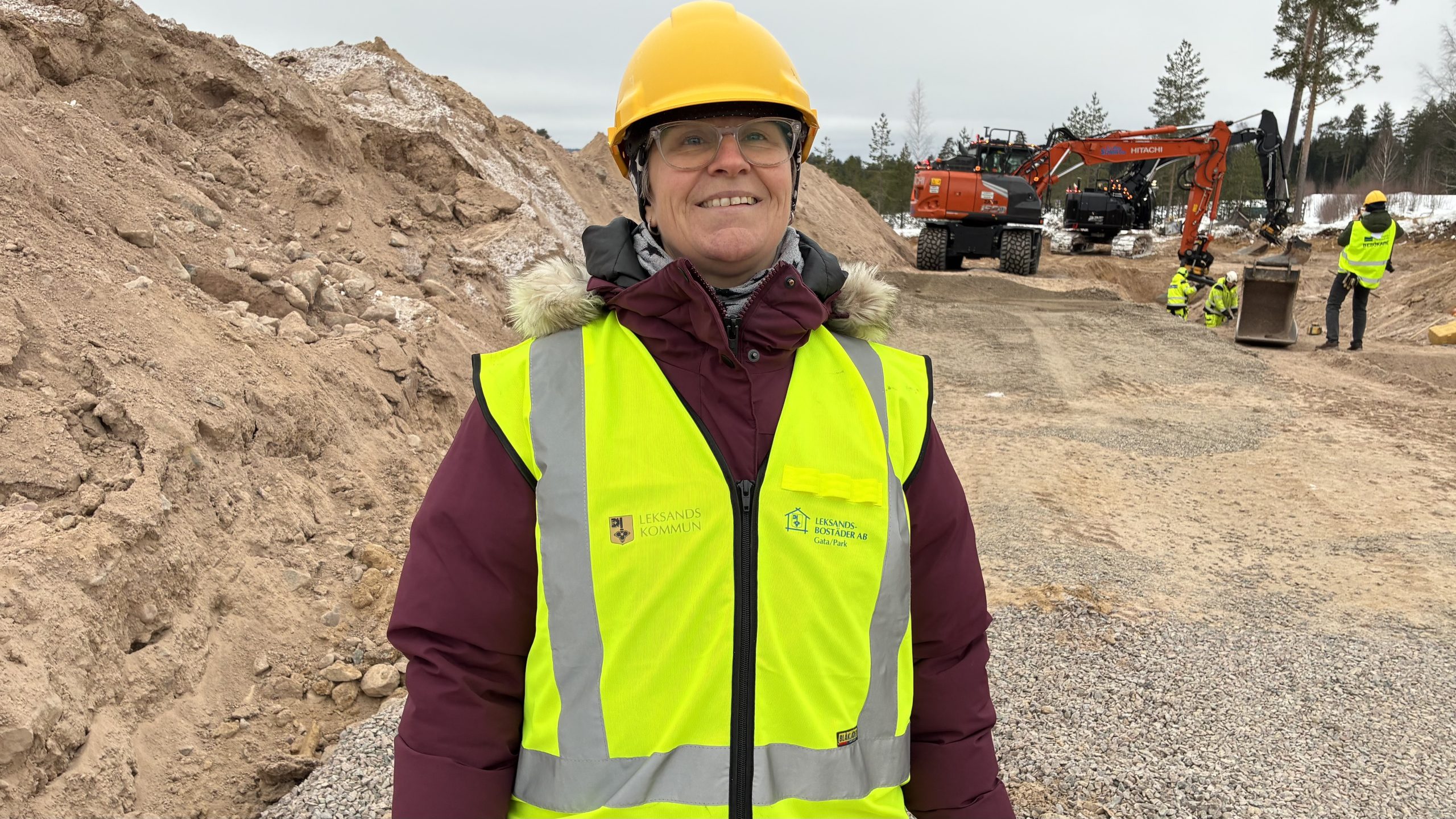 Kvinna med varselväst och hjälm framför grävmaskin på byggarbetsplats.