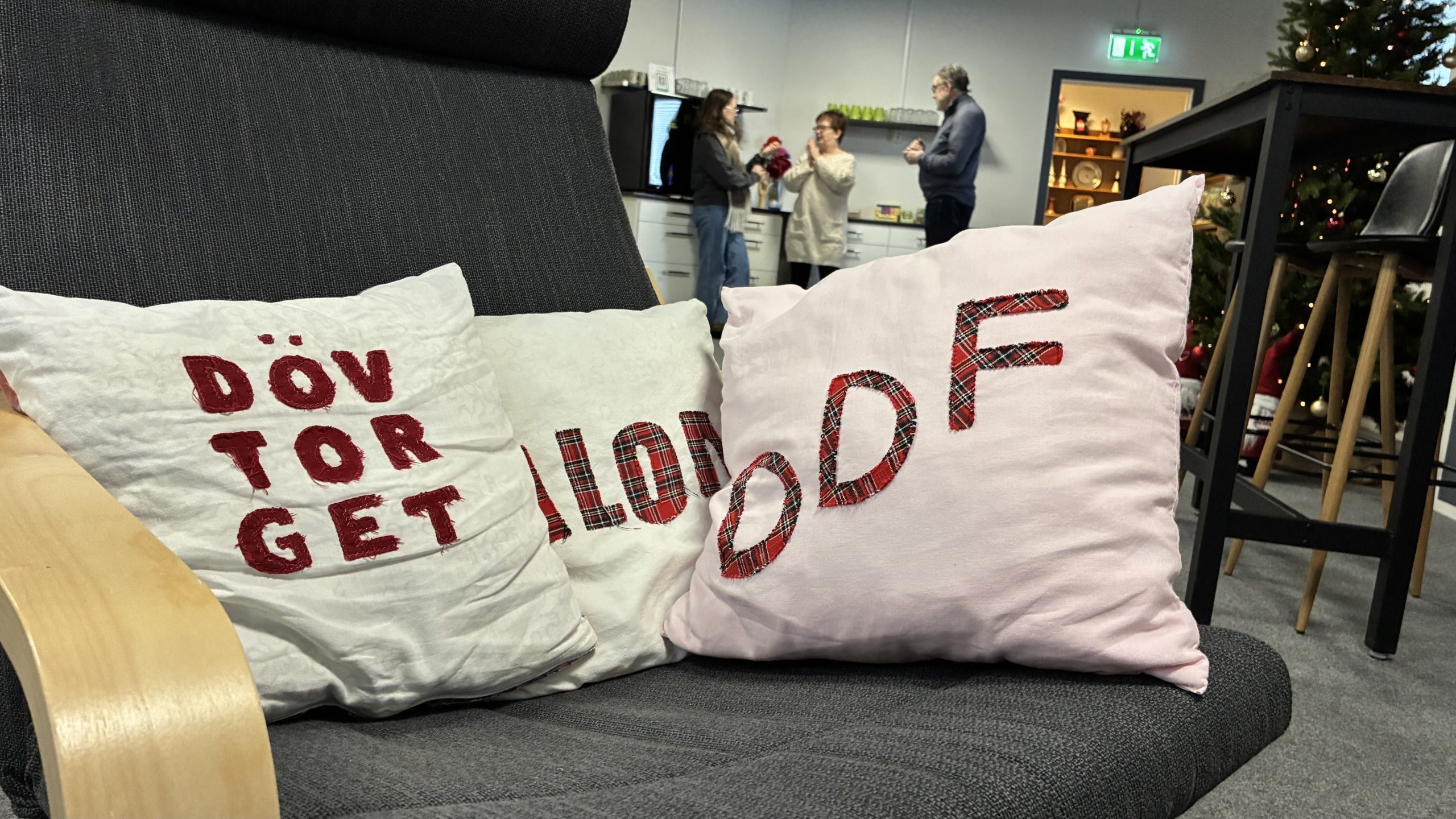 Närbild på kuddar med texten "Dövtorget" och "DDF". I bakgrunden står tre personer och tecknar med varandra.