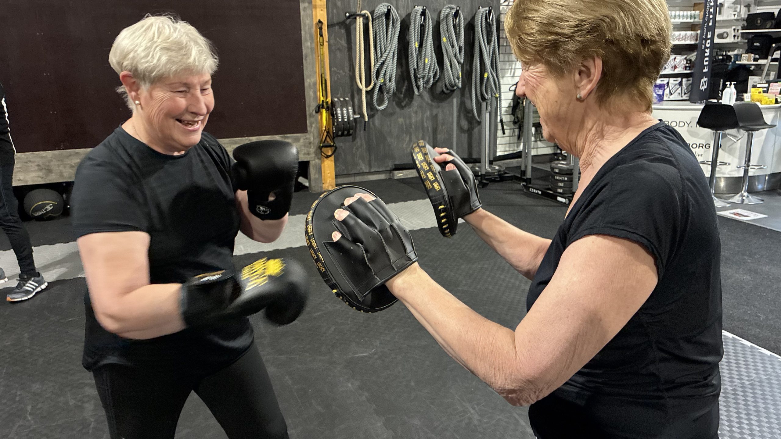 Två sprudlande glada äldre kvinnor står och tränar med boxningshanskar och slagkuddar.