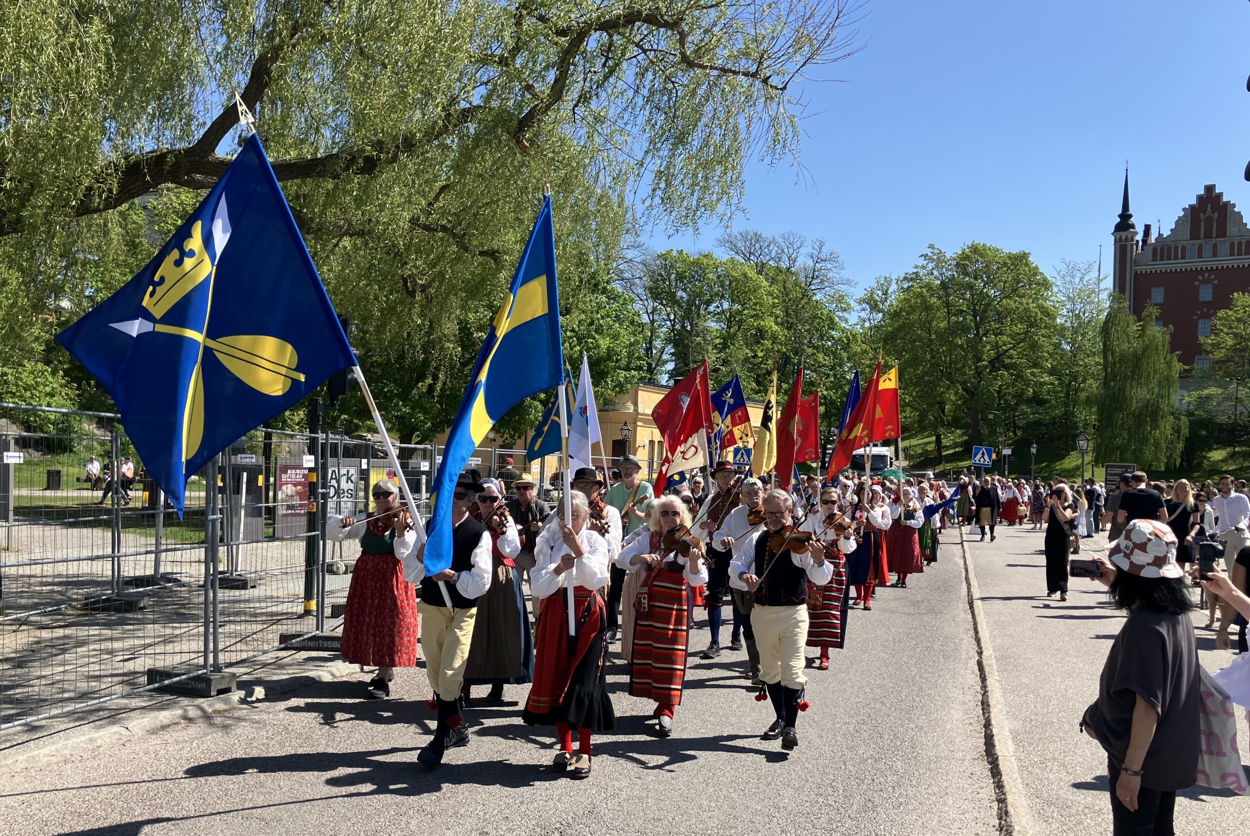 Långt tåg med personer i folkdräkt som håller upp flaggor, ackompanjerade av spelmän med fioler.