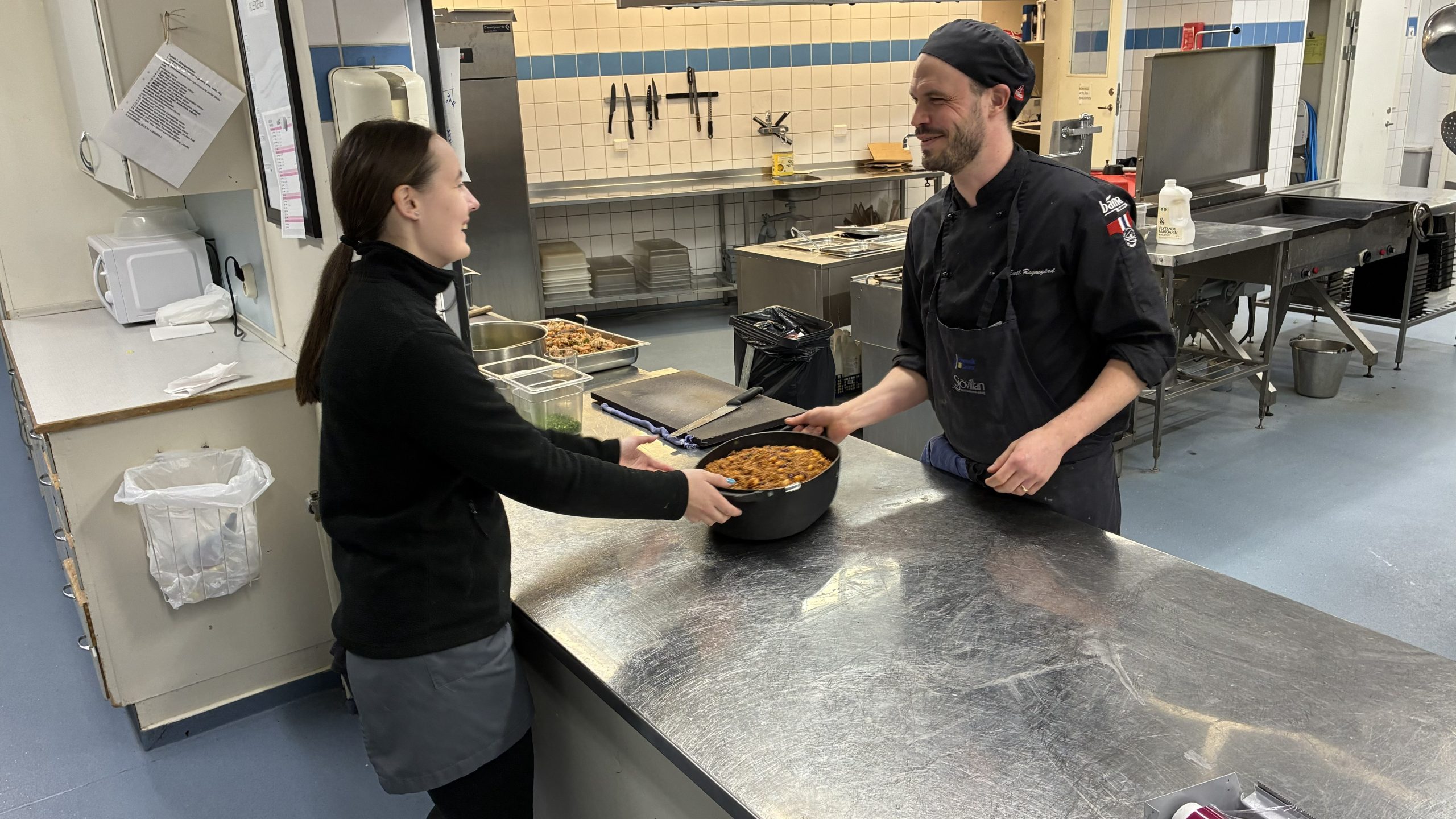 Två skrattande personer förbereder mat vid en serveringsdisk i köket.