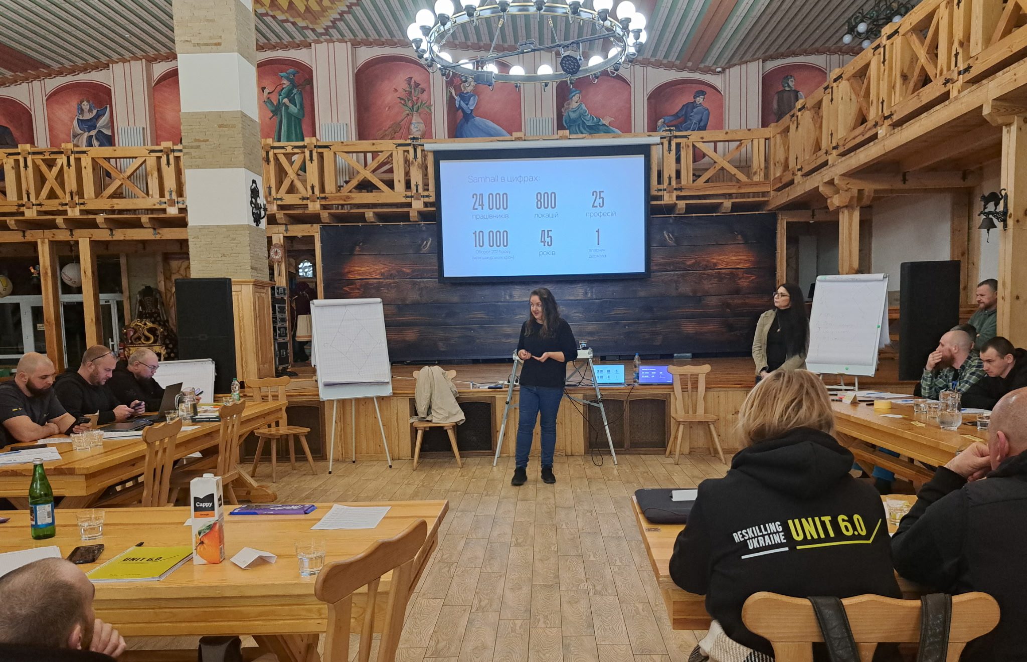 Kvinna står i pampig sal och föreläser framför en presentation, med deltagarna som lyssnar vid borden.