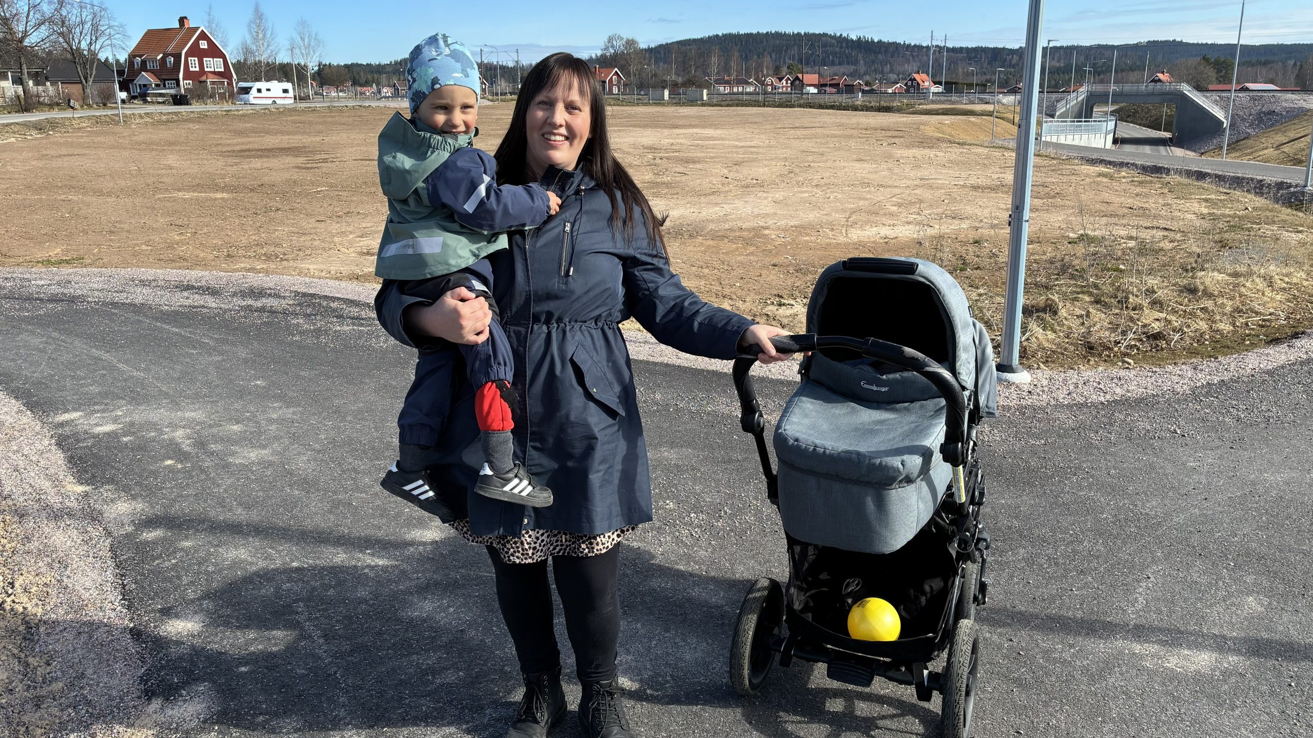 Kvinna håller barn i famnen och står bredvid en barnvagn framför en jordplätt intill järnvägen.