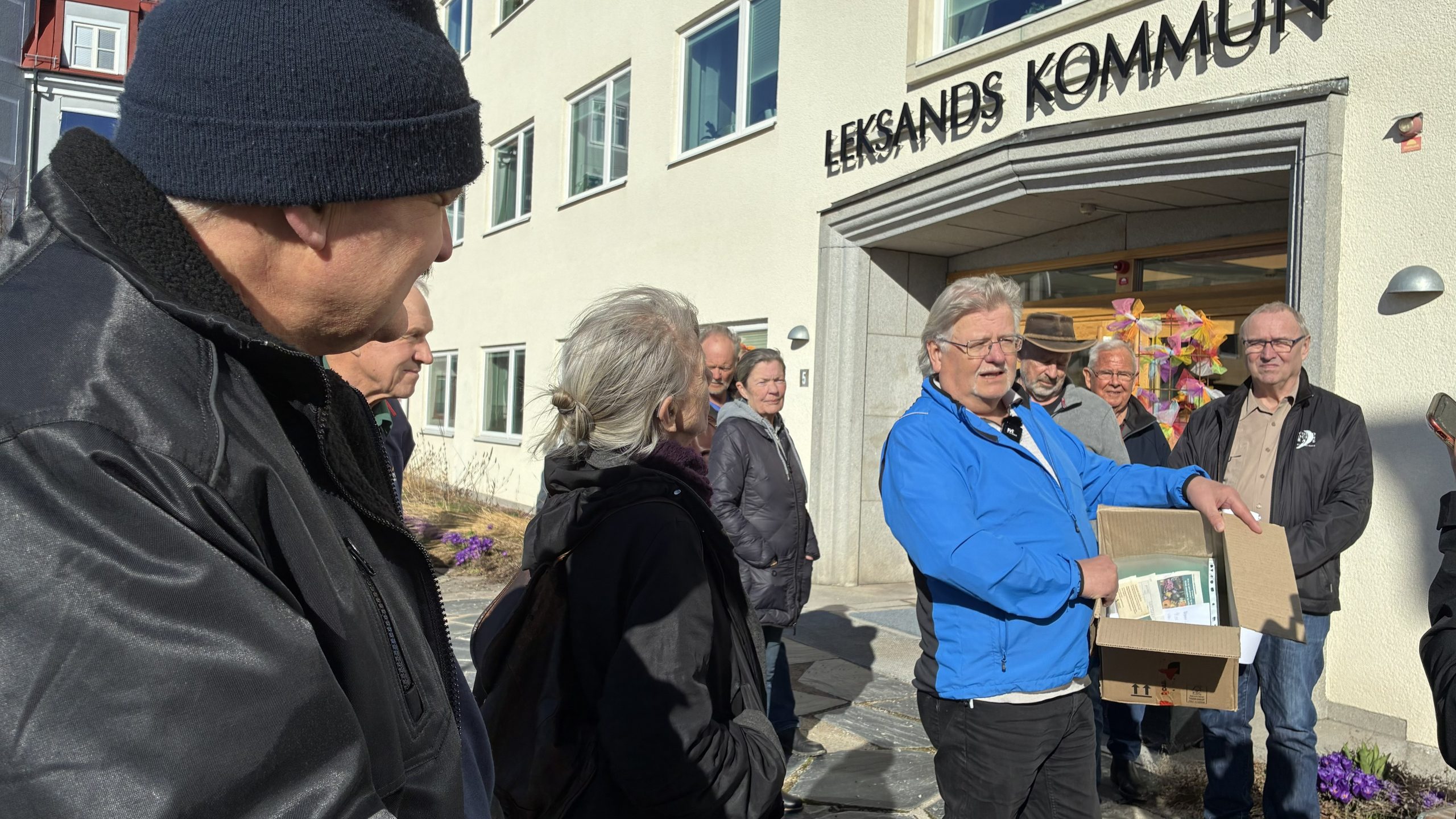 Grupp står i solsken utanför Leksands kommunhus. En man i mitten har en kartong innehållande papperslappar och tidningsurklipp.