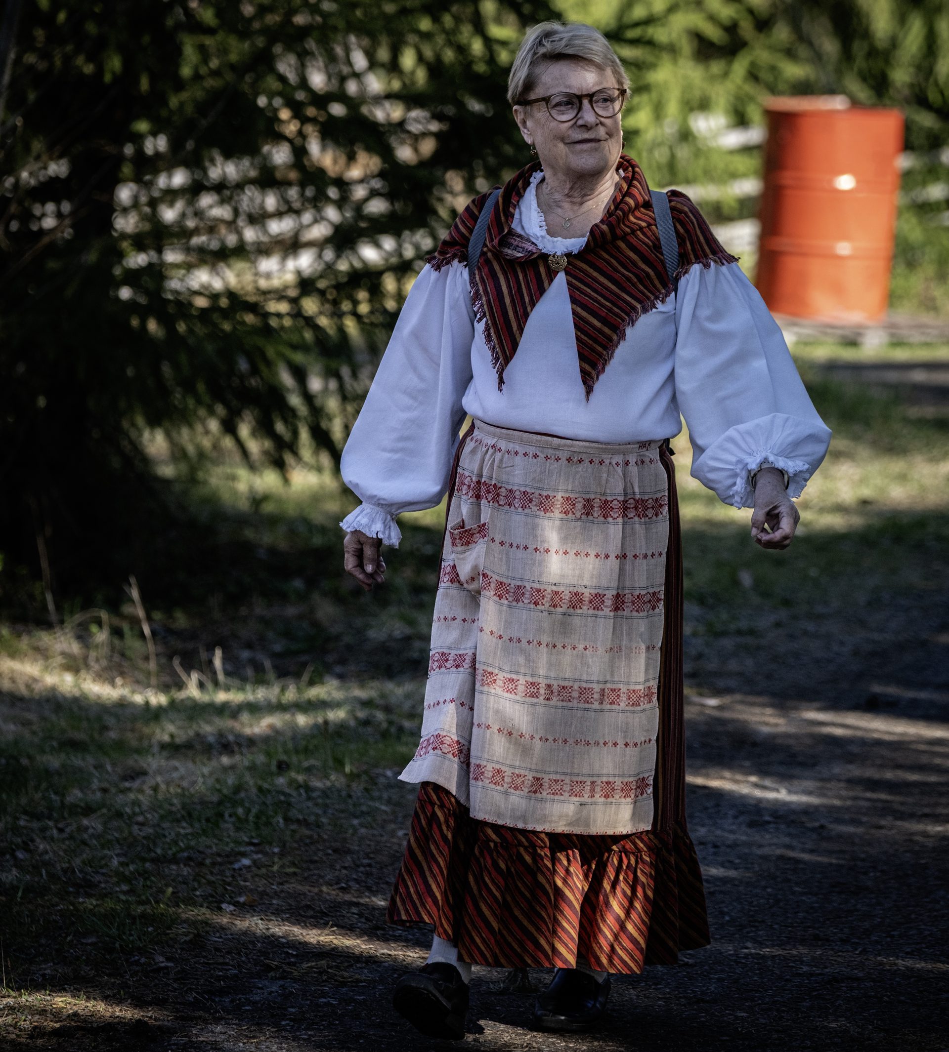 Kvinna i folkdräkt går på en väg med lite skog i bakgrunden.