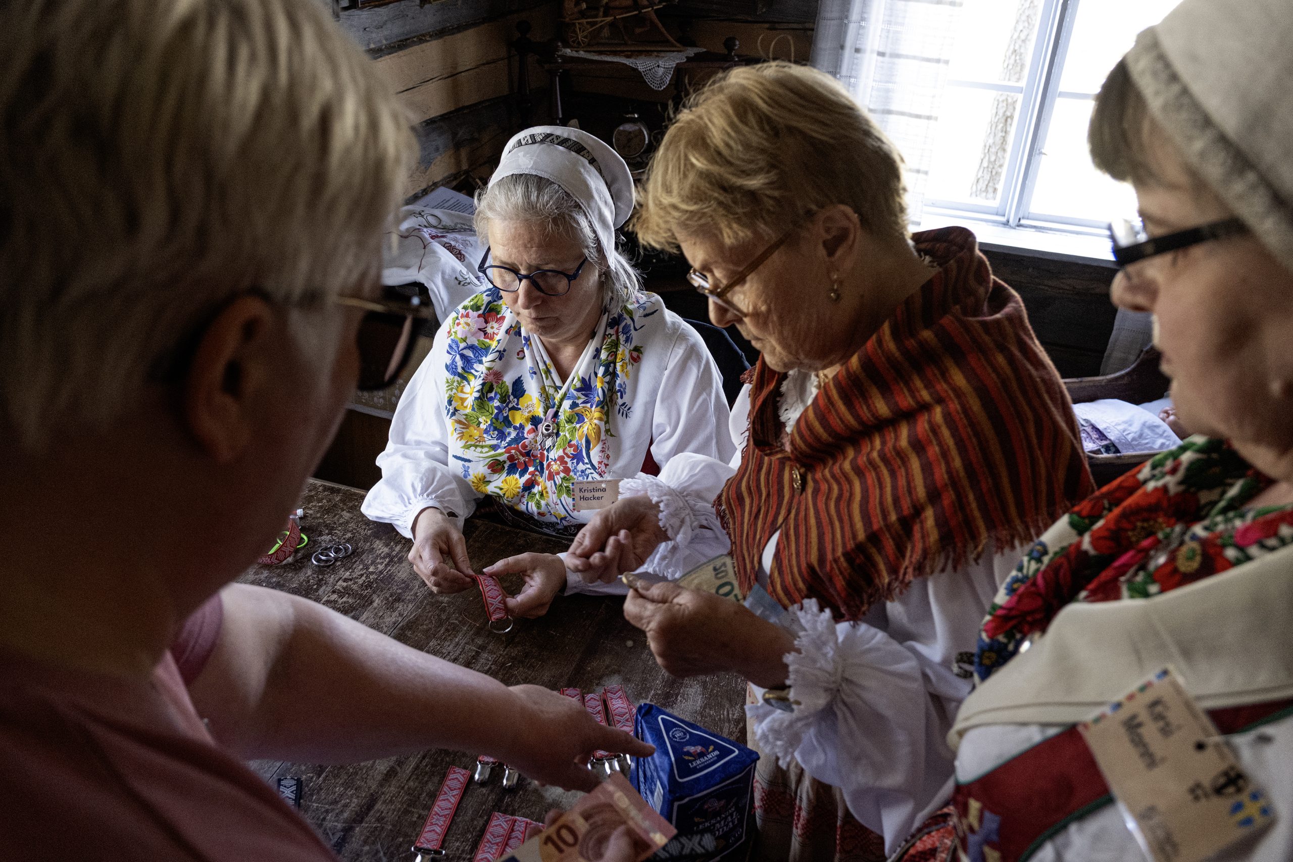 Kvinnor sträcker sedlar mellan varandra, med ett paket knäckebröd på bordet och några nyckelband med Leksands krusbands-motiv.