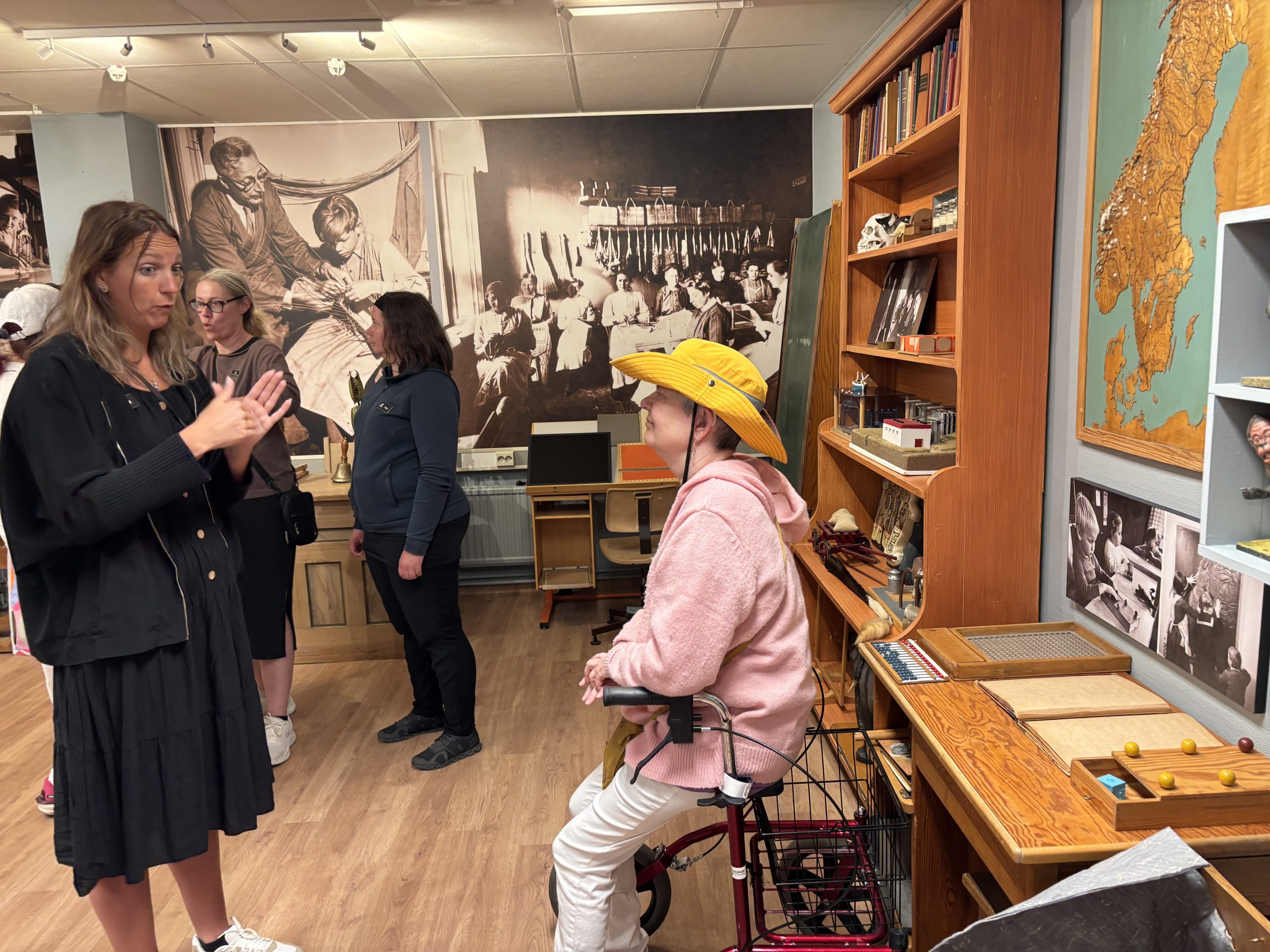 På väggen i museilokalerna hänger kartor och gamla bilder. Närmast kameran sitter Helena Karlsson lutad mot en rullator samtidigt som en tolk tecknar framför henne. I bakgrunden står fler personer och diskuterar.