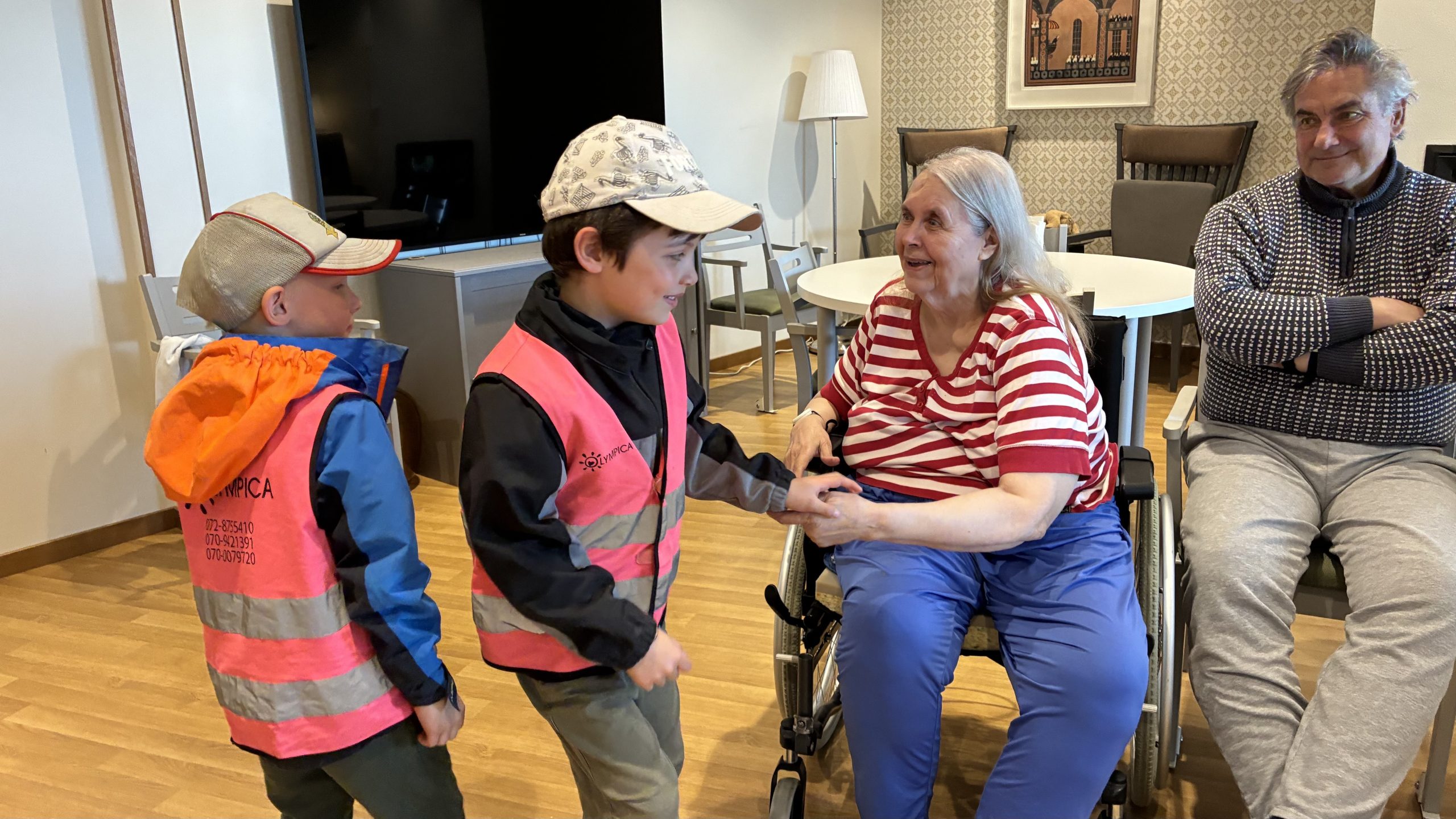 Två barn i reflexvästar skakar hand med en äldre kvinna som sitter i en stol.