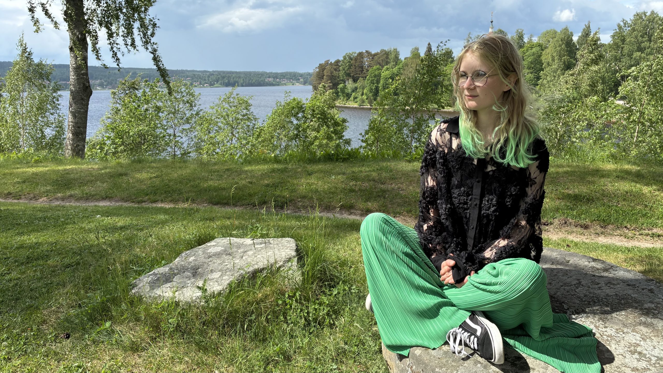 Ellen Hysing sitter med benen i kors på en sten med Siljan och Kyrkudden i Leksand i bakgrunden.