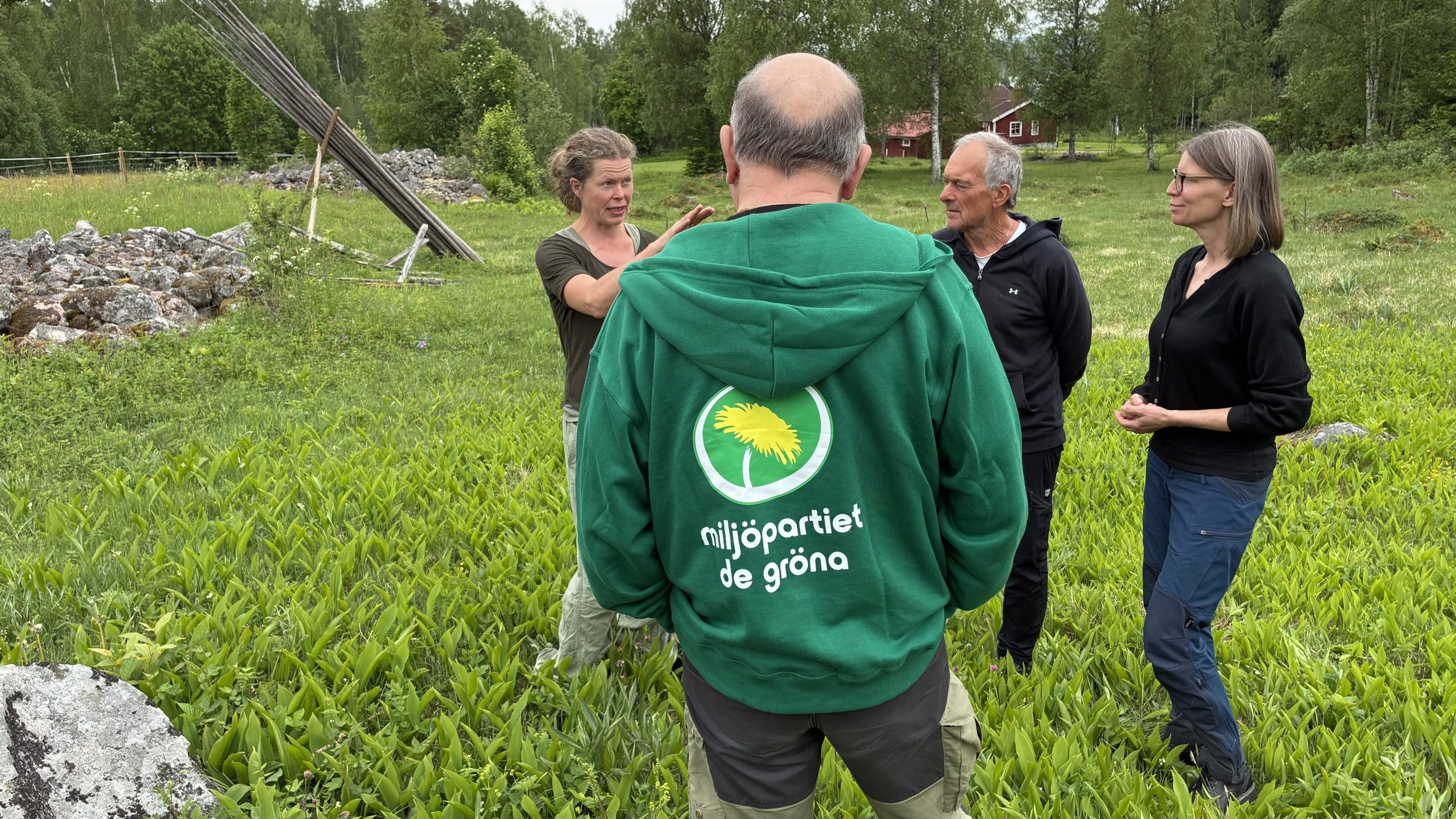 Olle Berglund står med ryggen vänd mot kameran och pratar med Göran Petri och andra partikamrater. Berglund visar ett stort tröjtryck med "Miljöpartiet de gröna" och maskrossymbolen