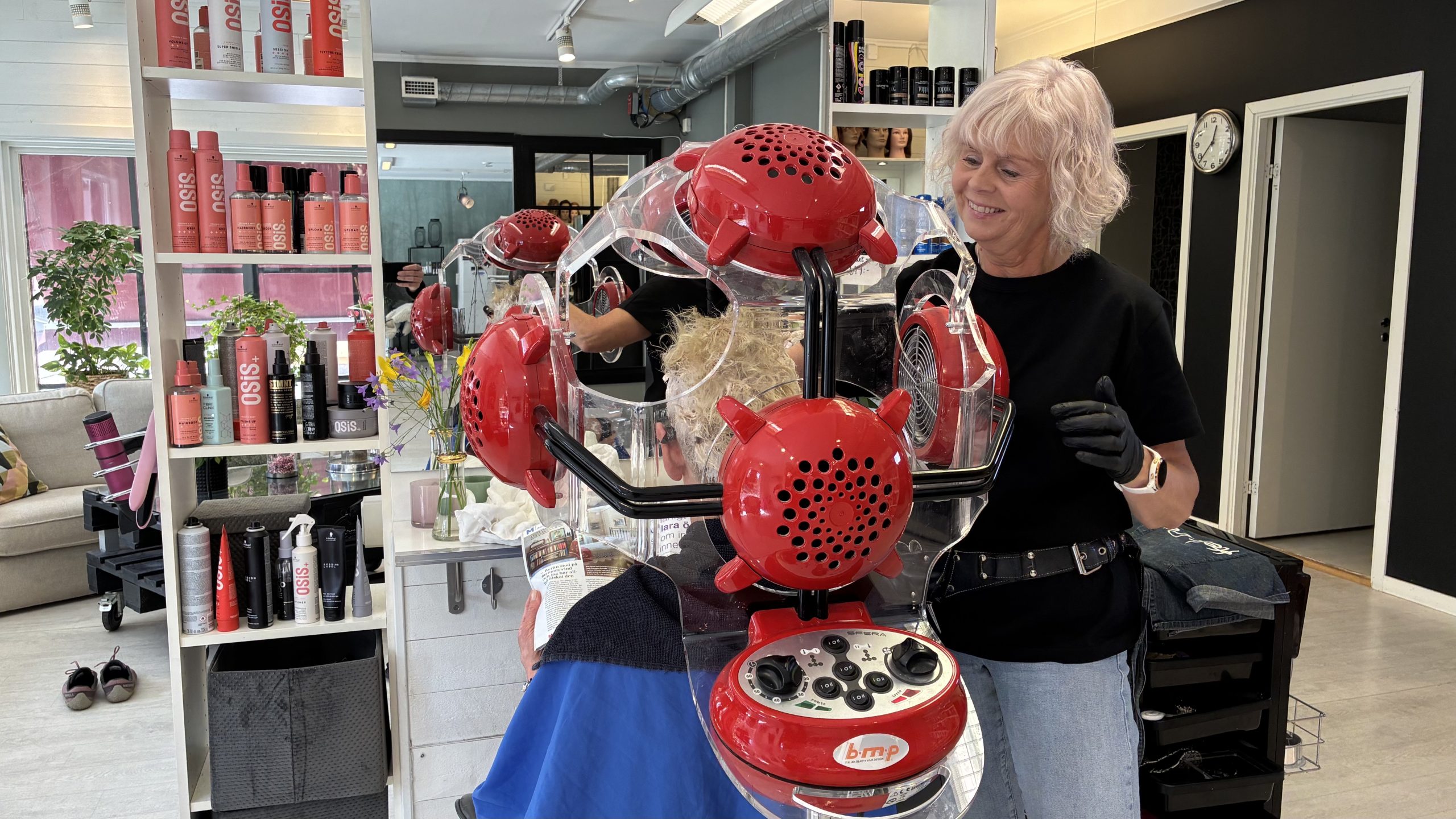 Leende kvinna klipper en kund som sitter med en stor röd apparat runt huvudet.