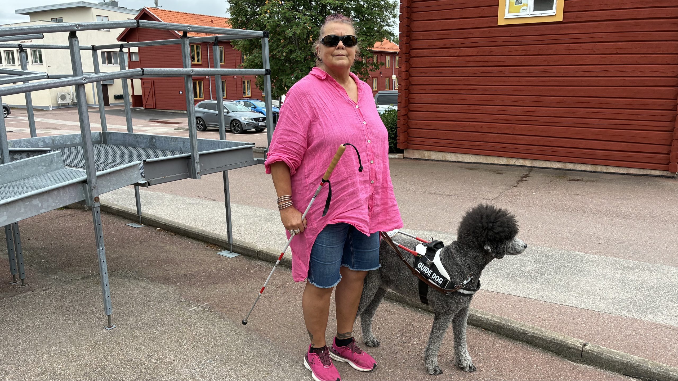 Kristina Nord står utomhus vid rampen till Dövtorget och håller ledarhundens handtag i den ena handen och käppen i den andra. Hunden är en hög grå pudel. Kristina ler och tittar mot kameran bakom sina solglasögon.