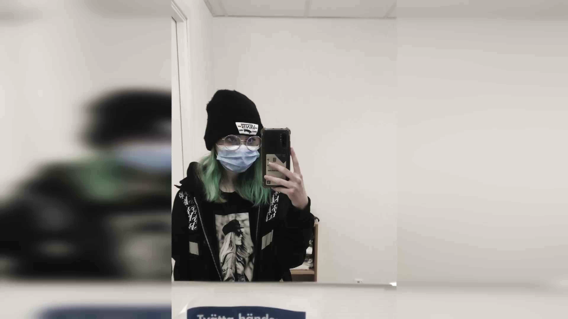Bild tagen med mobilkamera mot en spegel i sjukhusmiljö. Den unga kvinnan på bilden har munskydd och en mössa på sig, så att nästan bara glasögonen syns.