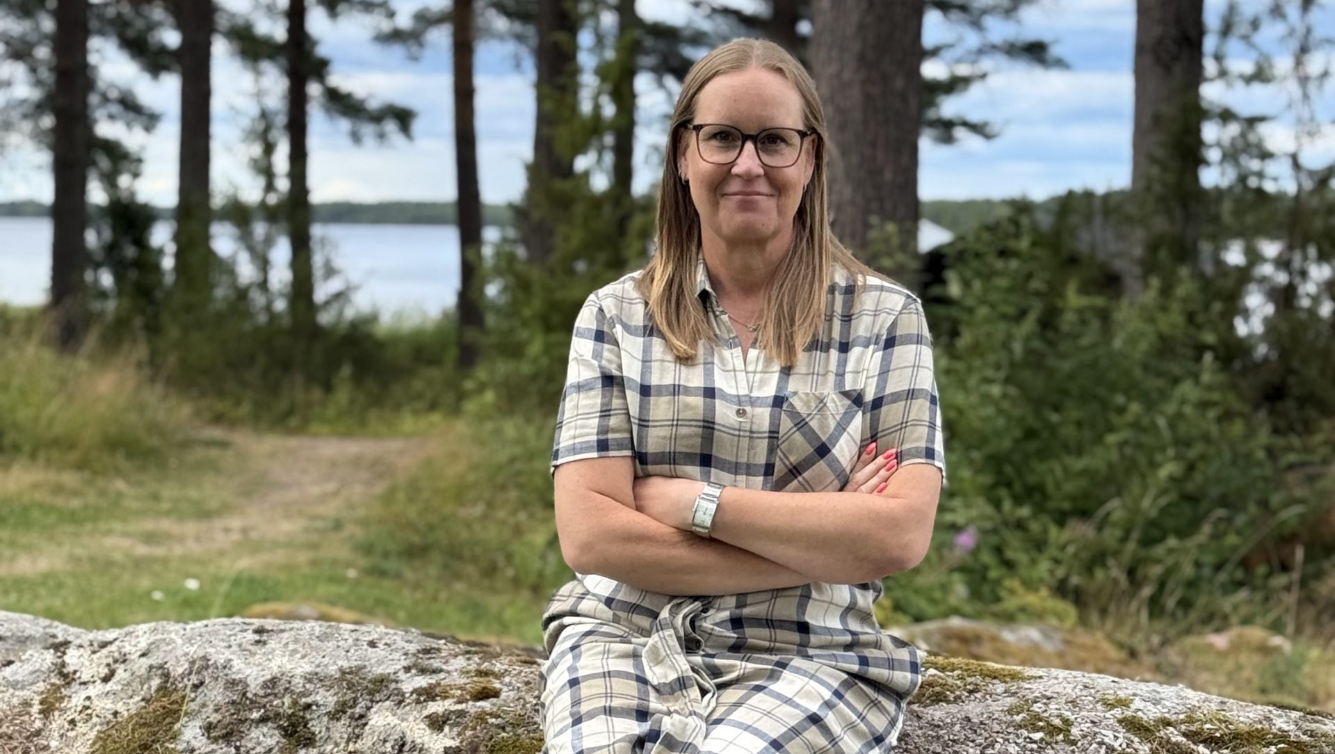 Karin Mikkonen sitter på en sten med korslagda armar, Siljan syns i bakgrunden.