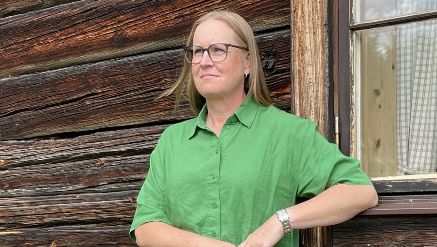 Karin Mikkonen står lutad mot en trähusfasad och blickar bort mot horisonten.