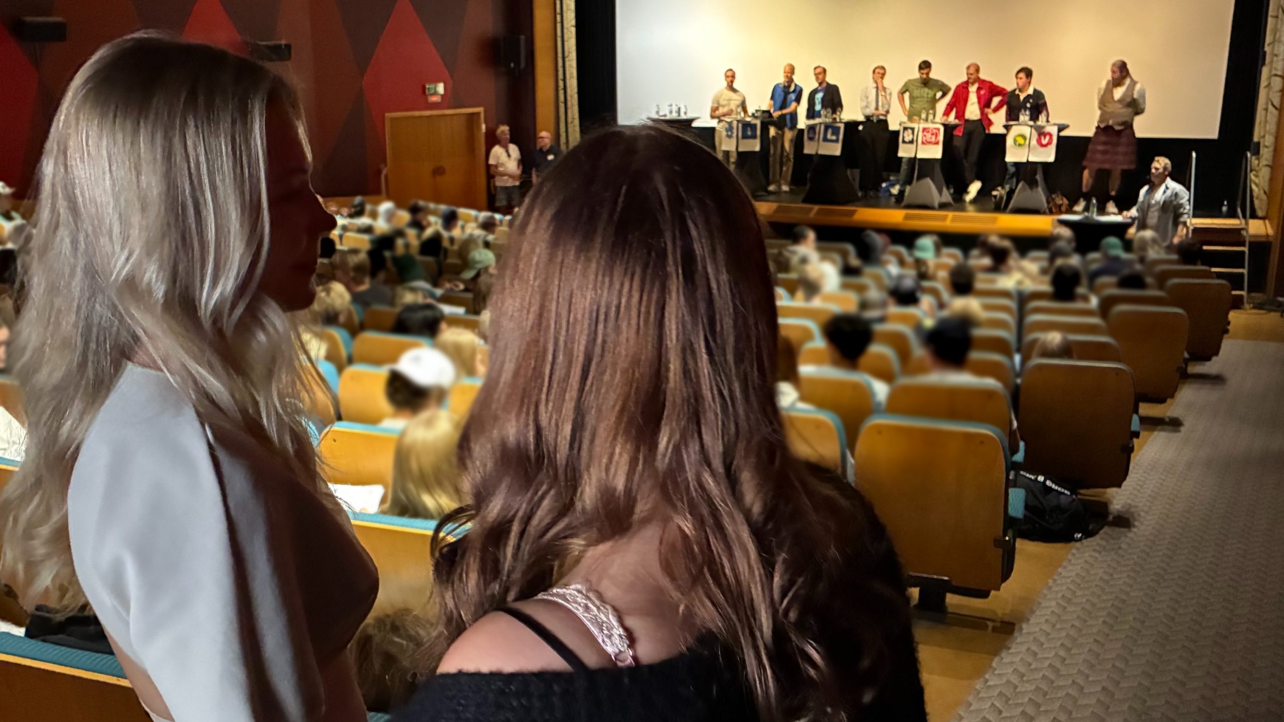 Två tonårstjejer tittar ut över en biosalong med politiker vid ståbord framme på scenen.