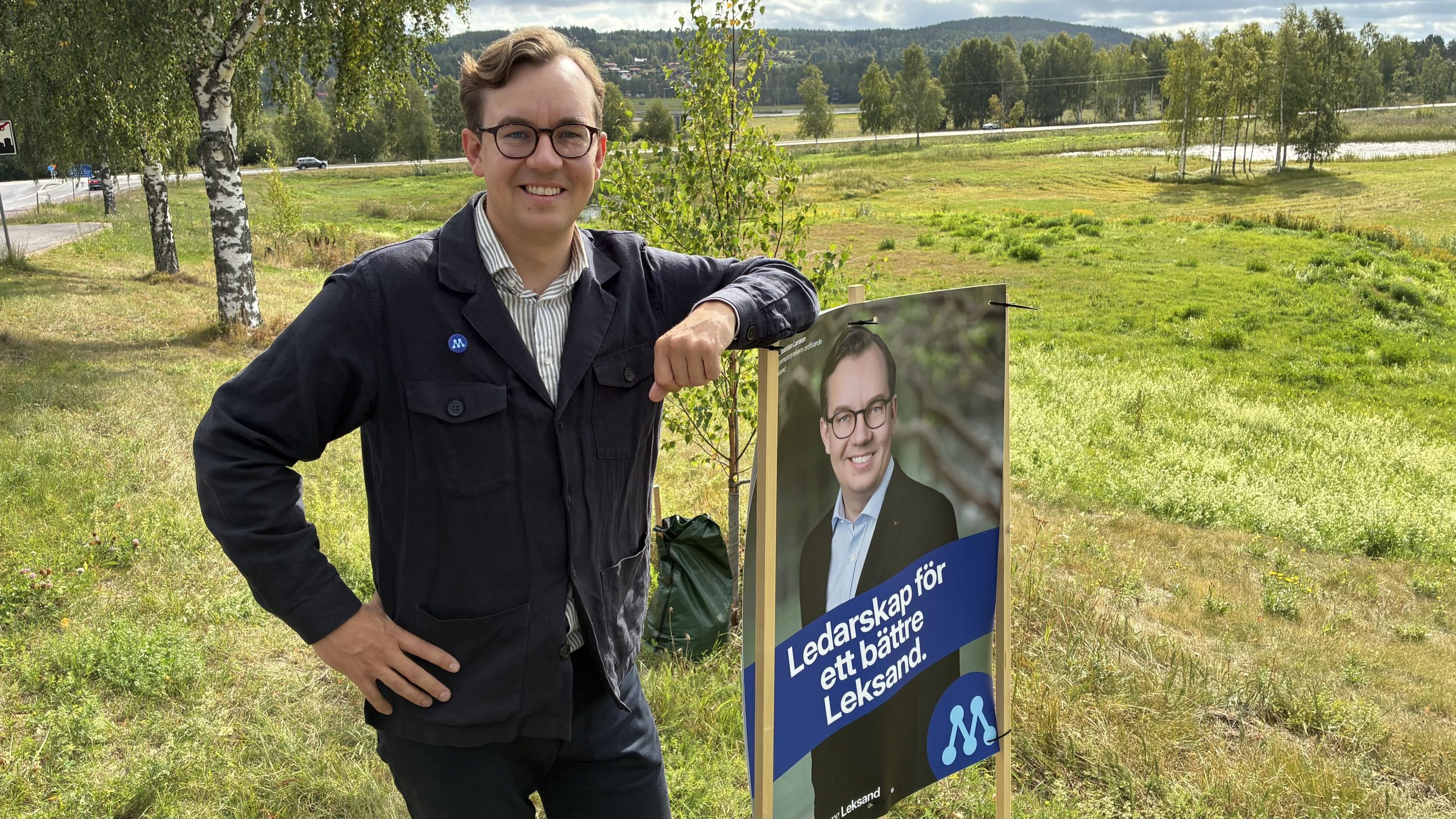 Sebastian Larsson står och poserar vid ett valplakat med hans ansikte och texten "Ledarskap för ett bättre Leksand".