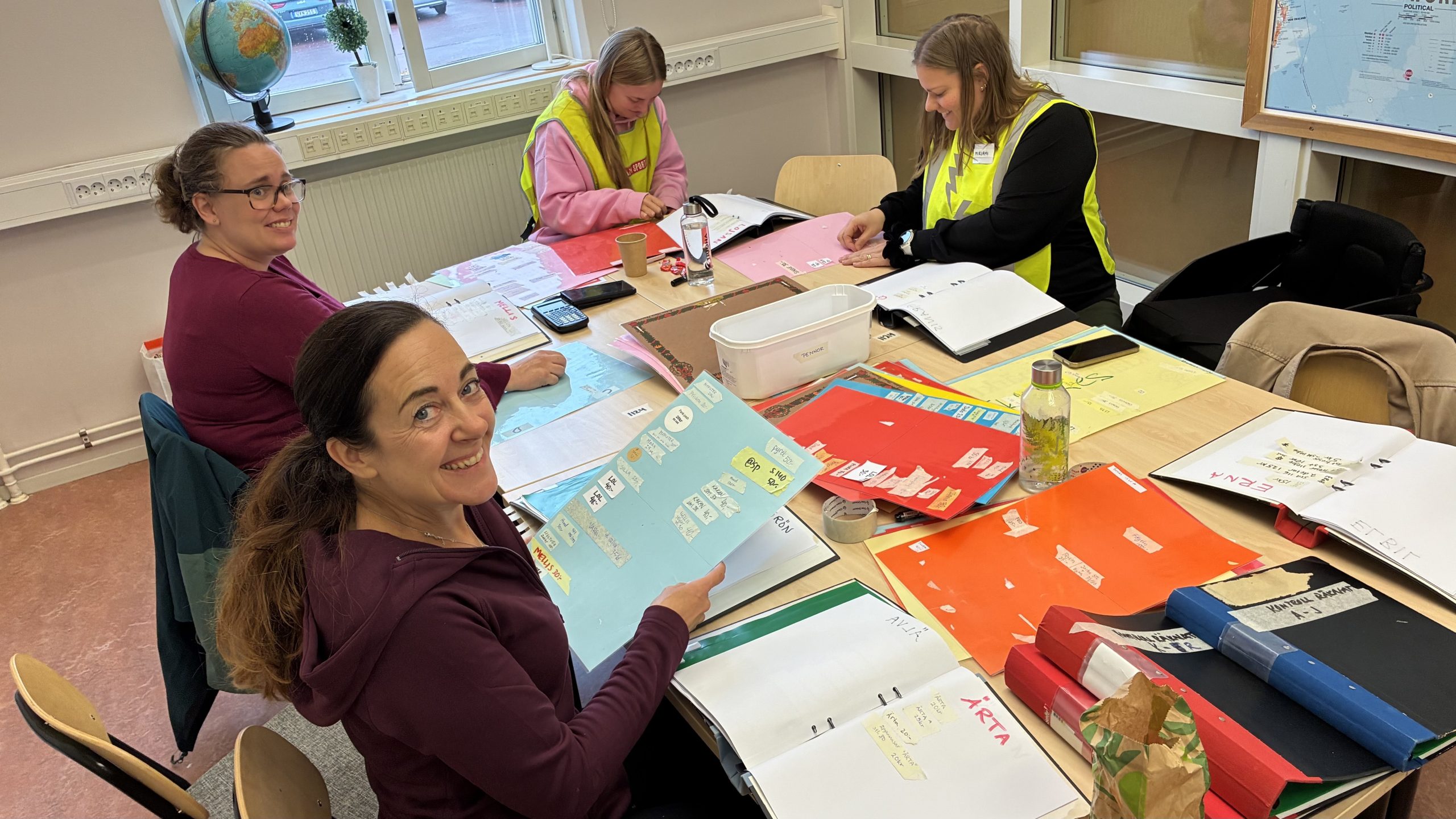 Fem personer sitter runt ett bord och sorterar tejplappar från olika brickor in i pärmar med ett alias för varje sida.