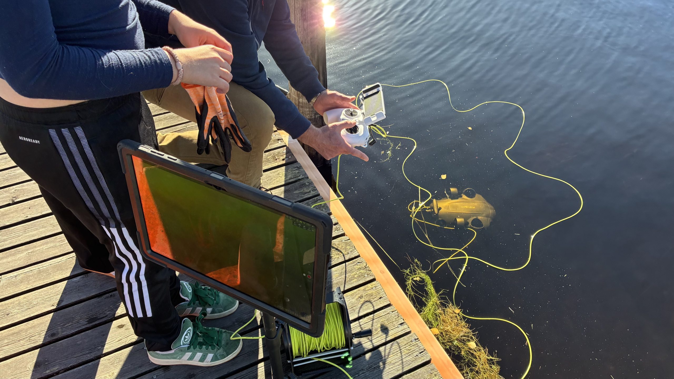 I vattnet syns en undervattensdrönare fäst med ett rep nära ytan, och på bryggkanten står några oidentifierbara personer med en handkontroll och en stor skärm.