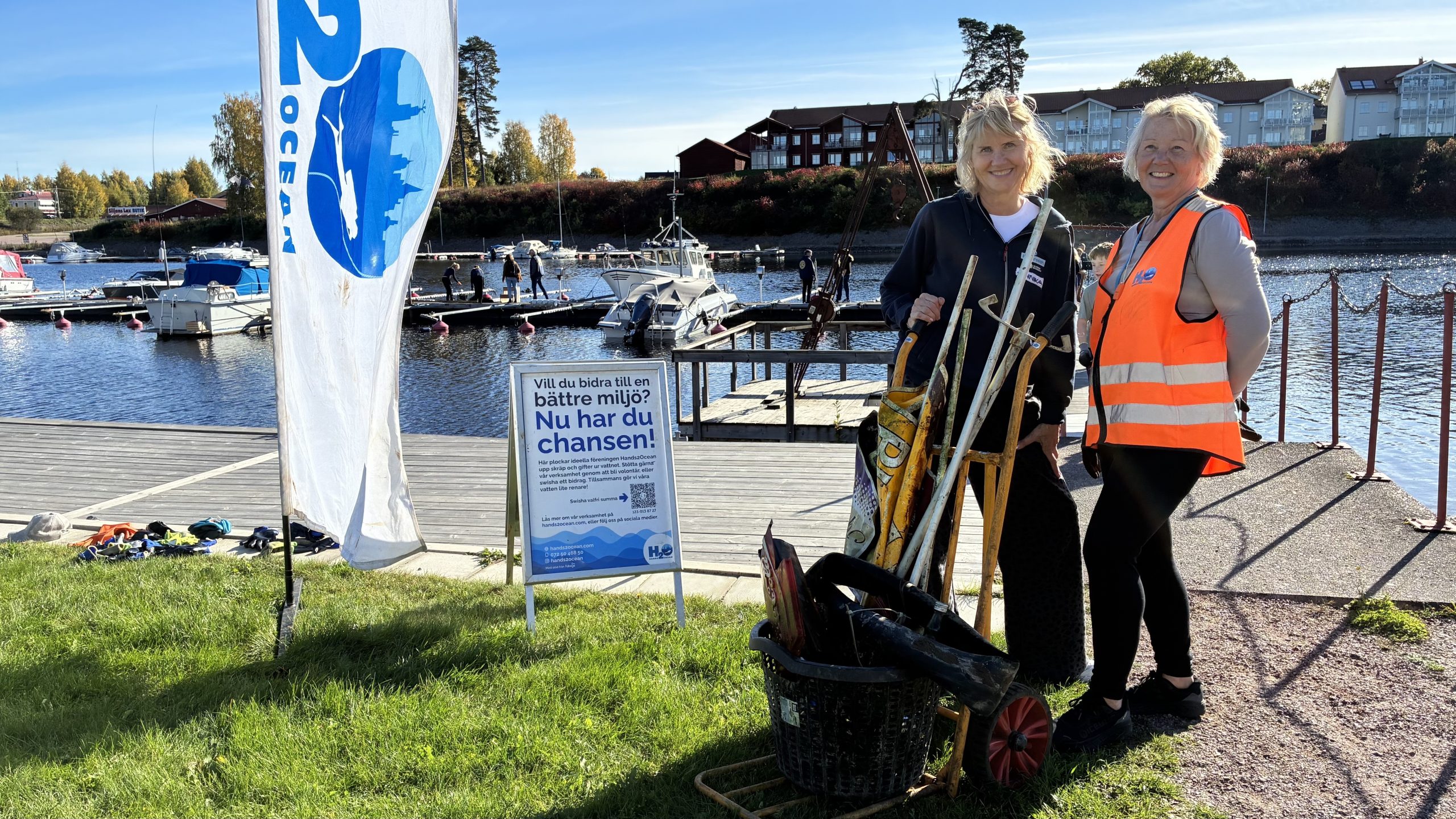 Två personer skjuter en pirra med älvskräp framför sig. I bakgrunden syns Hands2oceans marknadsföringsmaterial och bryggan vid kajen.