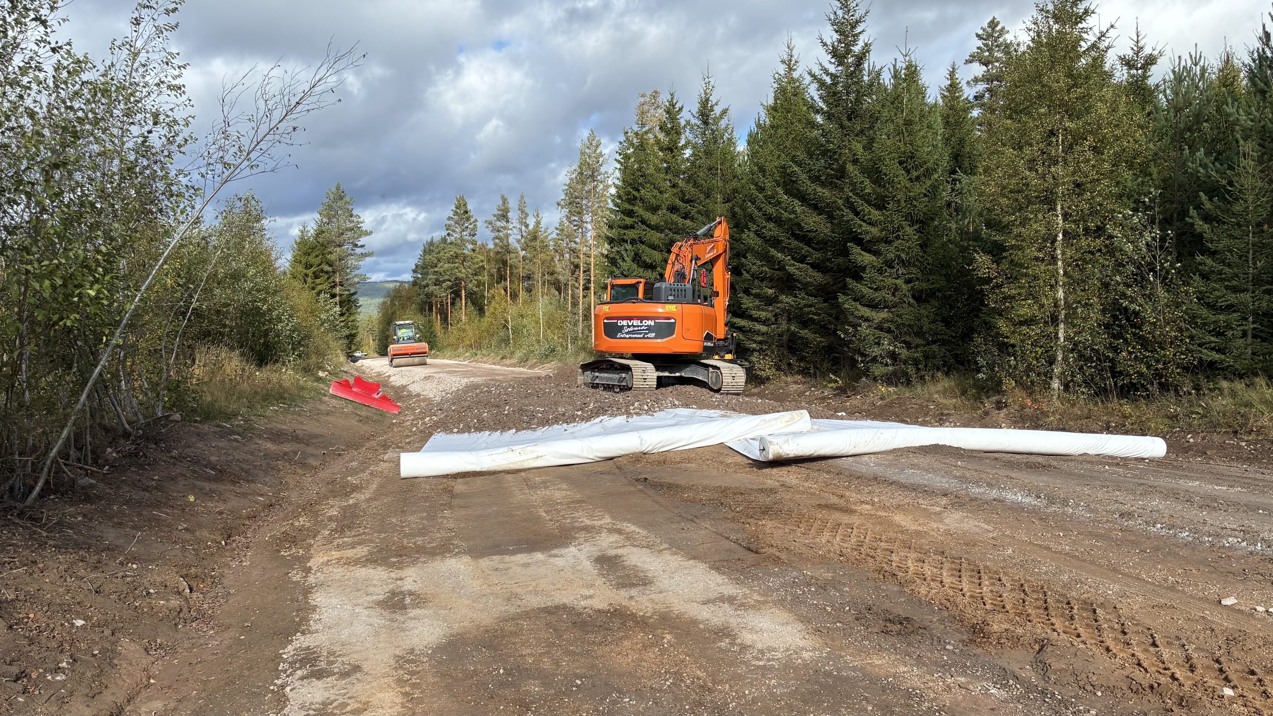 Stor grävmaskin rullar ut en duk och fördelar ut grus på en väg som ska breddas och förstärkas.