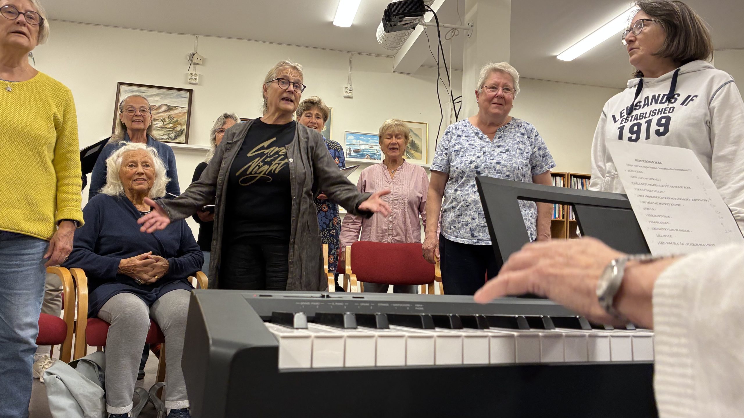 I förgrunden syns händer som spelar på ett keyboard, i bakgrunden kvinnor som sjunger med ett aktivt kroppsspråk.