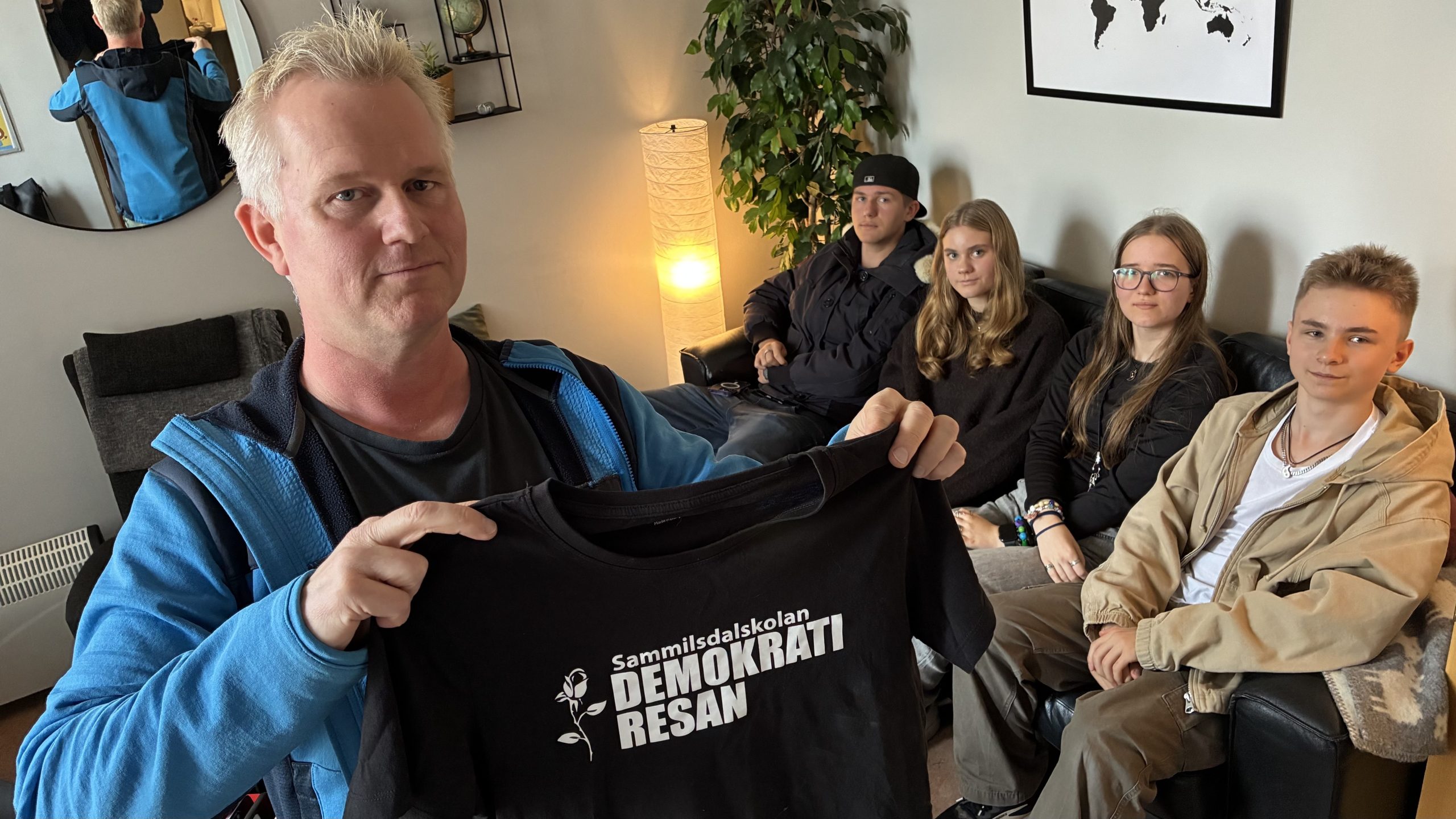 Stefan Olsson håller upp en svart t-shirt med vitt tryck: "Demokratiresan". I bakgrunden sitter fyra elever i soffan.