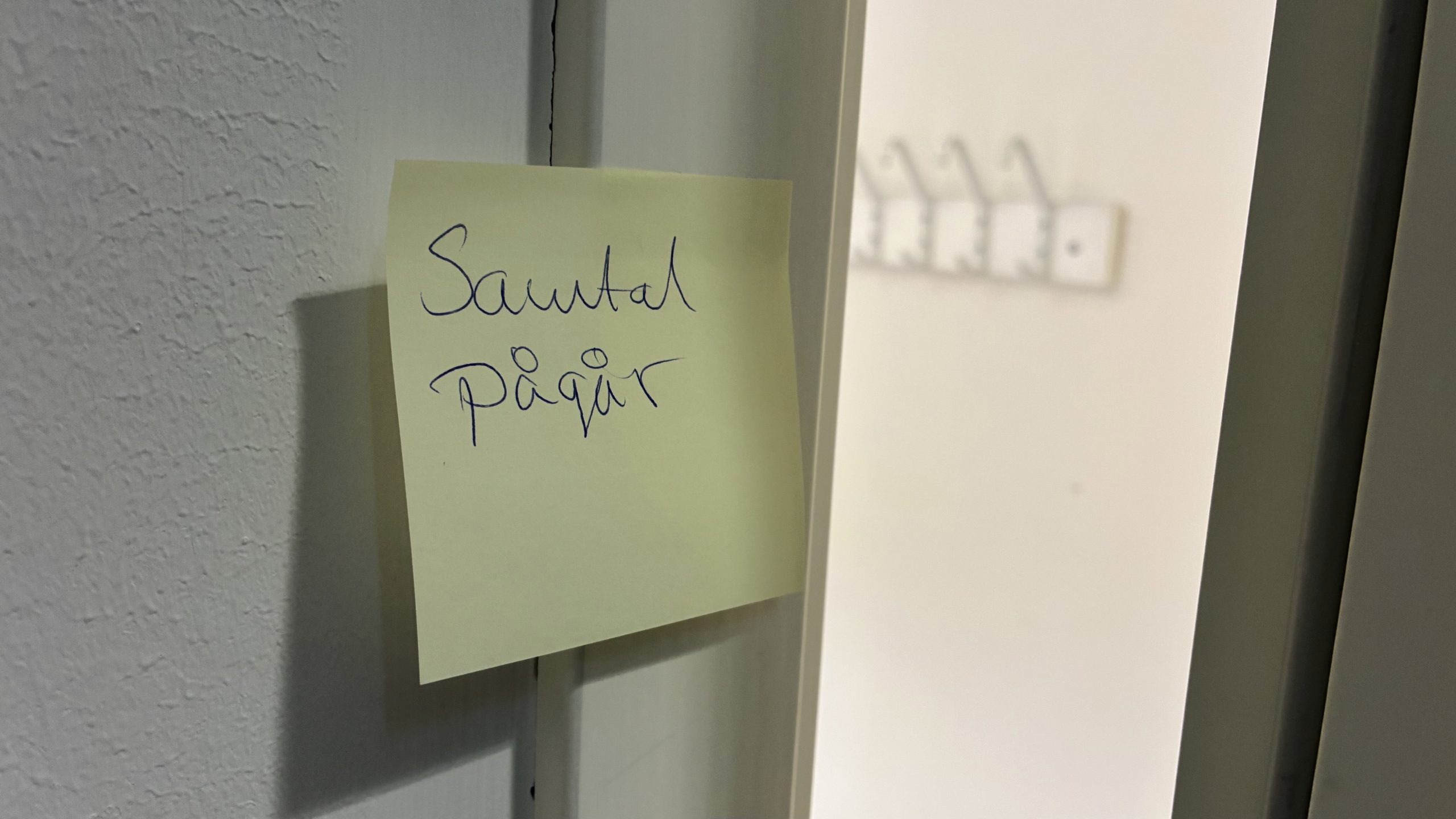 Bild på post-it lapp med texten "Samtal pågår", vid en dörr som står på glänt. På rummets insida syns några klädhängare.