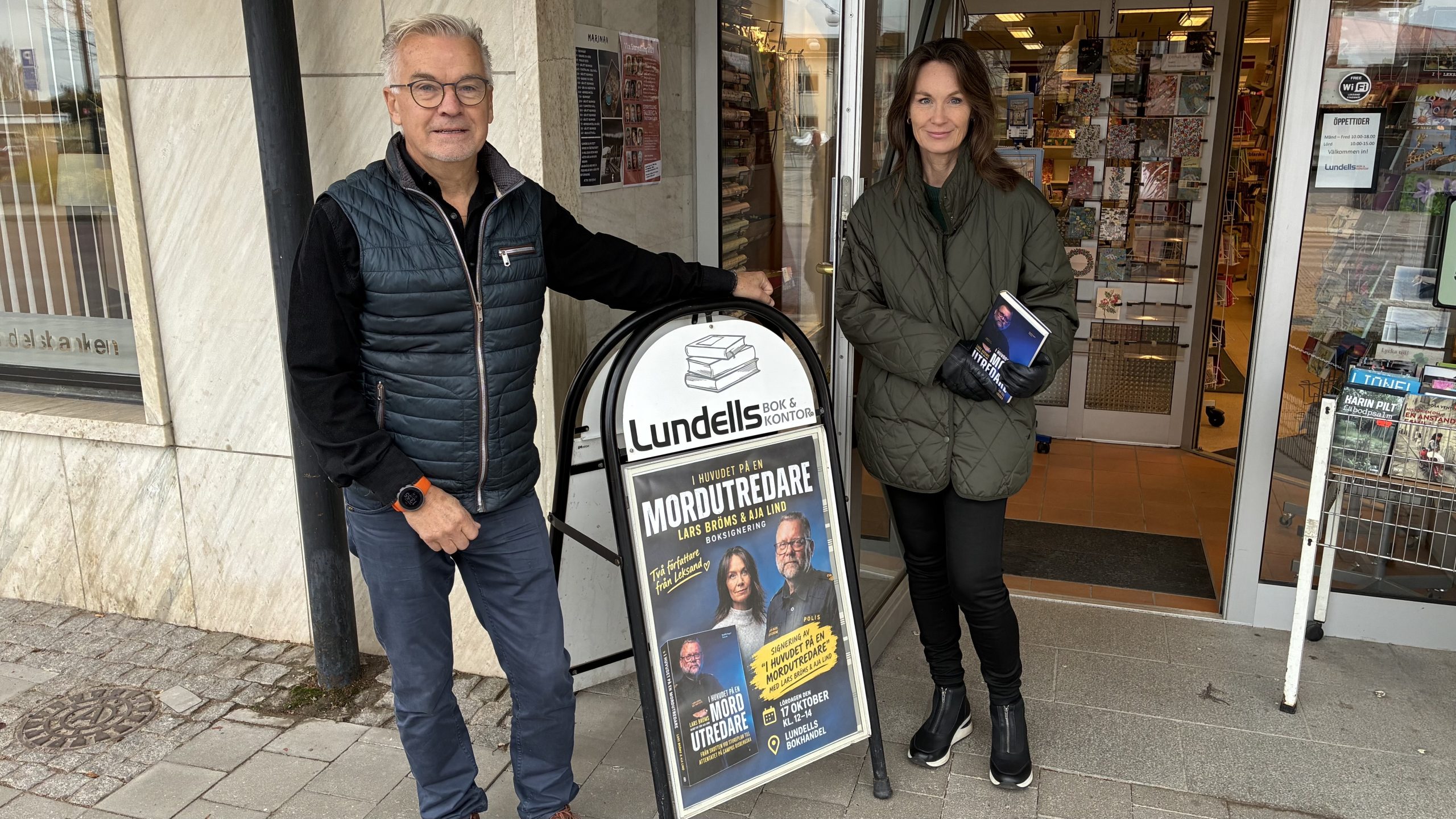Göran Lundell och Aja Lind utanför bokhandelns entré med ett affischställ mellan dem som bjuder in till boksigneringen.