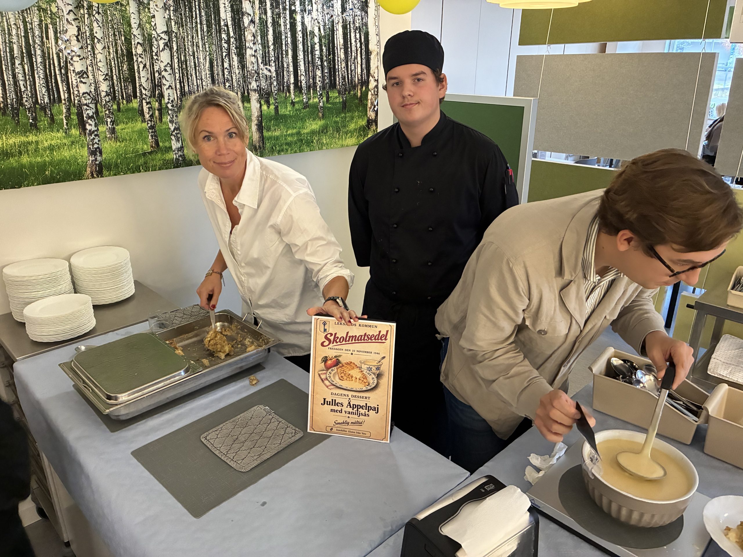 Tre personer står och serverar äppelpaj med vaniljsås i en skolmatsal