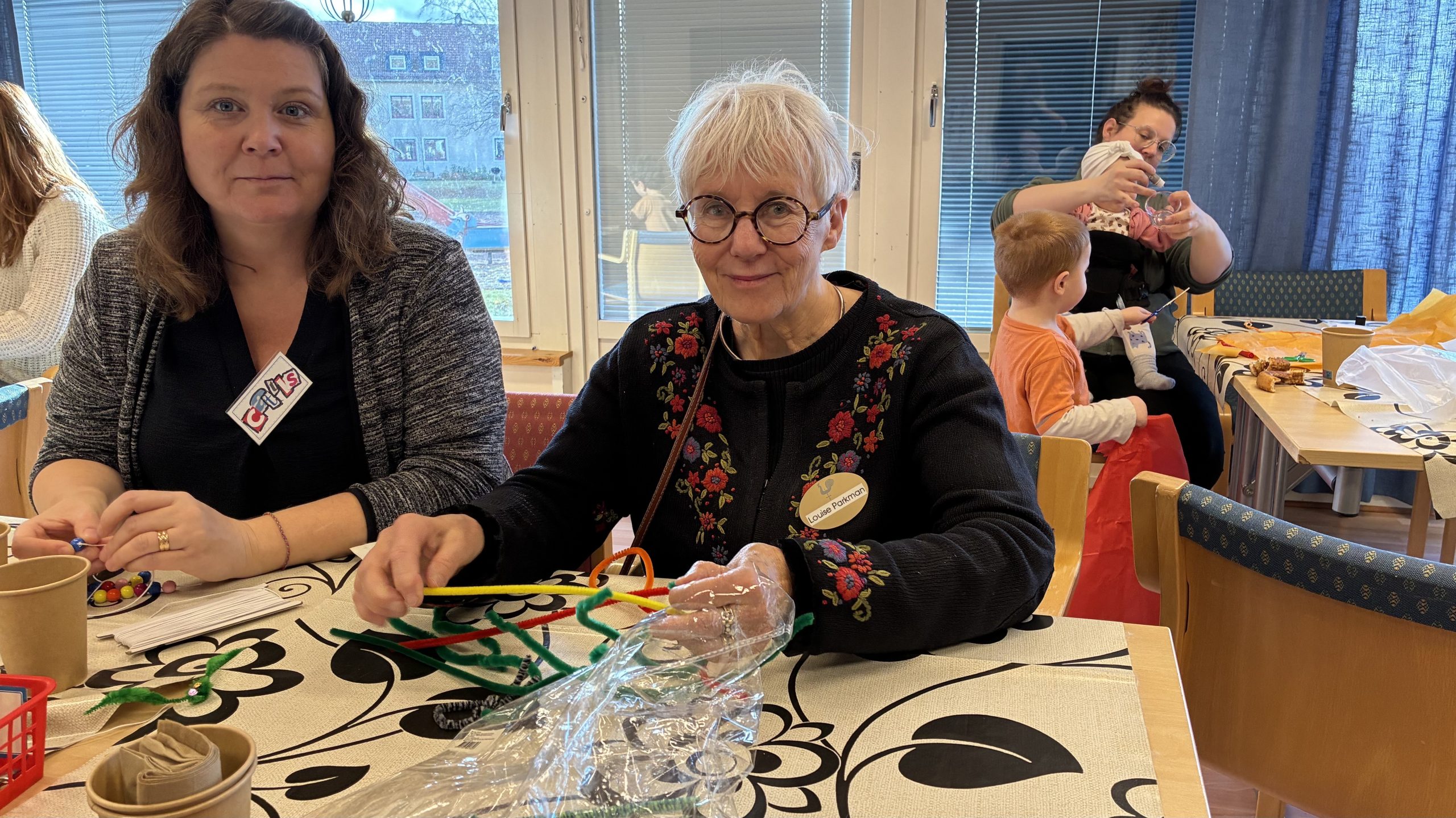 Två kvinnor sitter och pysslar med piprensare, med barnfamiljer i bakgrunden.