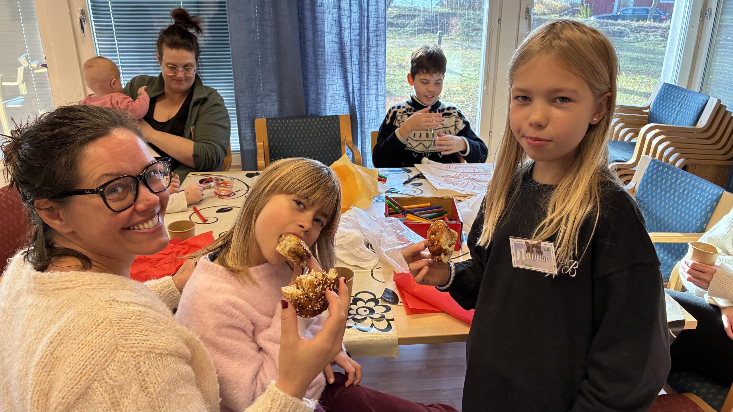 Mamma med flera barn sitter runt ett pysselbord och äter kanelbullar.
