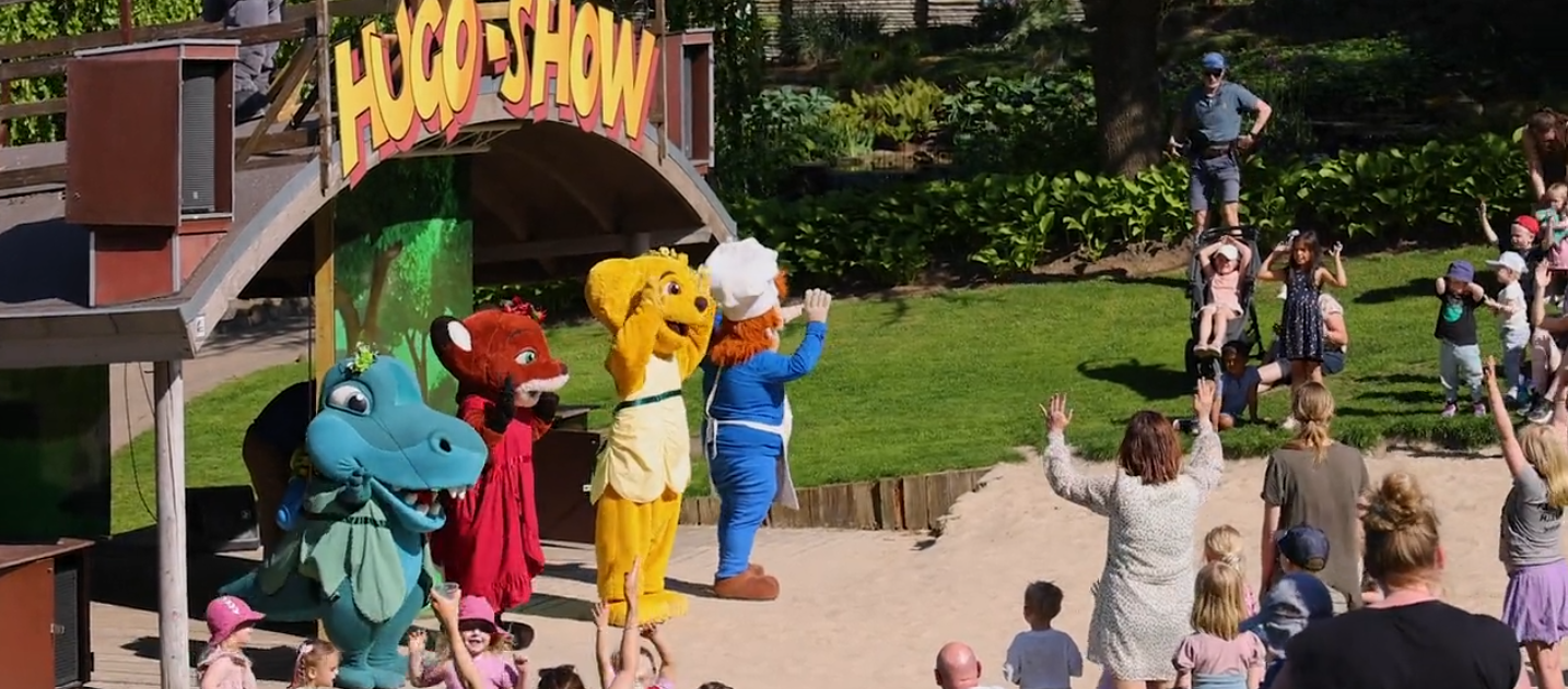 En gul björn tillsammans med några andra karaktärer showar framför barnfamiljerna. Ovanför scenen syns en stor rubrik: "Hugo Show".