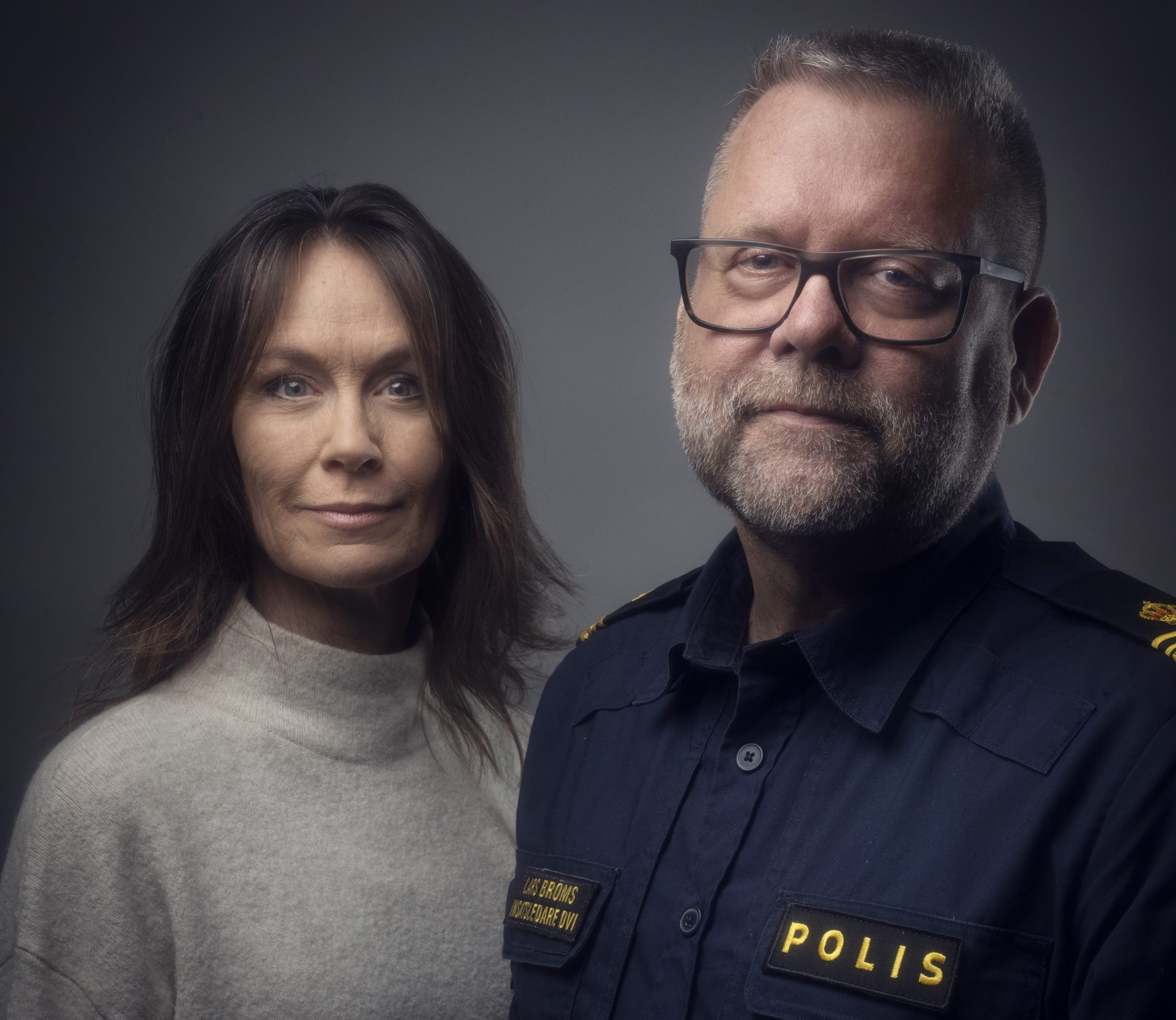Pressbild med grå bakgrund där Aja Lind och Lars Bröms står uppställda framför kameran i snyggt ljus. Aja i grå polotröja, Lars i polisuniform med "Insatsledare DVI" på bröstetiketten.